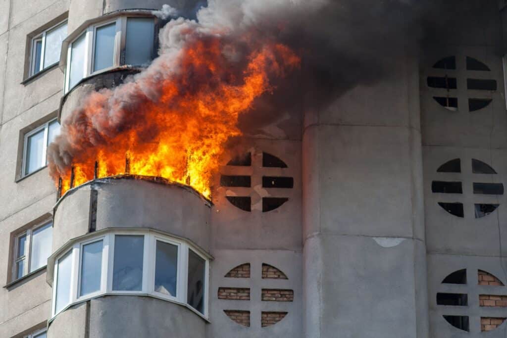 Außenansicht eines Mehrfamilienhauses. Eine Wohnung brennt, Flammen schlagen aus den Fenstern.